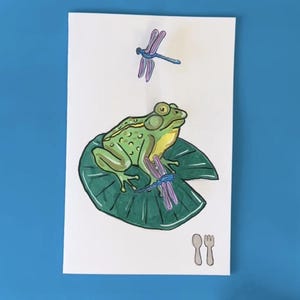 Puede incluir: Una ilustración dibujada a mano de una rana verde sentada en una hoja de nenúfar con dos libélulas azules. La rana tiene ojos grandes y negros y una barriga amarilla. La hoja de nenúfar es verde y tiene un contorno blanco. Las libélulas son azules y tienen contornos negros. La ilustración está sobre un fondo blanco. El texto "@moonshotkidz" está escrito debajo de la ilustración.