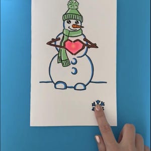 May include: A hand-drawn illustration of a snowman wearing a green hat and scarf, holding a heart. The snowman has a carrot nose and twig arms. A snowflake is drawn at the bottom. The text "@moonshotkidz" is at the bottom.