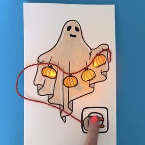 May include: A hand-drawn illustration of a ghost with a string of pumpkin lights. The ghost is beige with a black outline and a simple face. The string of lights is red, and the pumpkins are orange. The text "@moonshotkidz" is at the top and bottom.