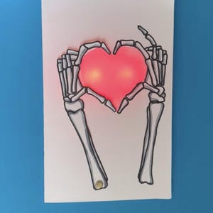 Pode incluir: Um cartão branco com um desenho de linha preta de duas mãos esqueléticas formando um coração em torno de um coração vermelho com uma luz dentro. O texto "@moonshotkidz" está na parte inferior do cartão.