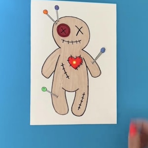 May include: A hand-drawn voodoo doll with a red heart in the center, surrounded by pins with colored heads. The doll is drawn on a white card with a brown outline. The background is a bright blue surface. The text "@moonshotkidz" is visible at the bottom of the image.