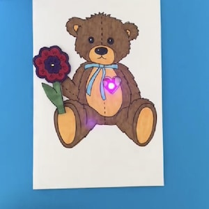 May include: A handmade greeting card with a brown teddy bear holding a red flower. The bear has a blue ribbon around its neck and a pink heart on its chest that lights up.