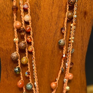 May include: A multi-strand necklace featuring a peach-colored woven cord. The necklace is adorned with a variety of beads in shades of brown, orange, green, and clear crystals. The beads vary in size and texture, creating a unique and bohemian style.