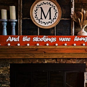 Puede incluir: Un letrero de madera rojo con la frase "And the stockings were hung" en escritura blanca, adornado con copos de nieve y clavijas blancas para colgar calcetines. El letrero se exhibe sobre una chimenea, con velas y una corona decorativa en el fondo.