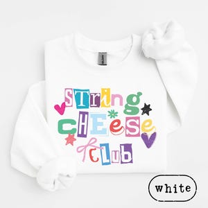 Könnte beinhalten: Weißes Sweatshirt mit dem Text "String Cheese Club" in bunten Buchstaben im Ausschnitt-Stil. Das Design umfasst Herzen, Sterne und eine Schleife. Das Sweatshirt hat lange Ärmel und einen Rundhalsausschnitt.