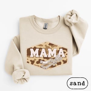 Könnte beinhalten: Sandfarbenes Sweatshirt mit dem Wort "MAMA" in weißen Blockbuchstaben in einem braunen und beigefarbenen Tarn-Sechseck. Darunter eine Grafik eines gebündelten Objekts. Das Wort "sand" steht unter dem Sweatshirt.