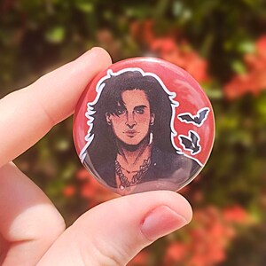 May include: A round button with a red background features a black and white illustration of a man with long black hair. He is wearing a black leather jacket and a silver necklace. Two black bats are flying in the upper right corner of the button.