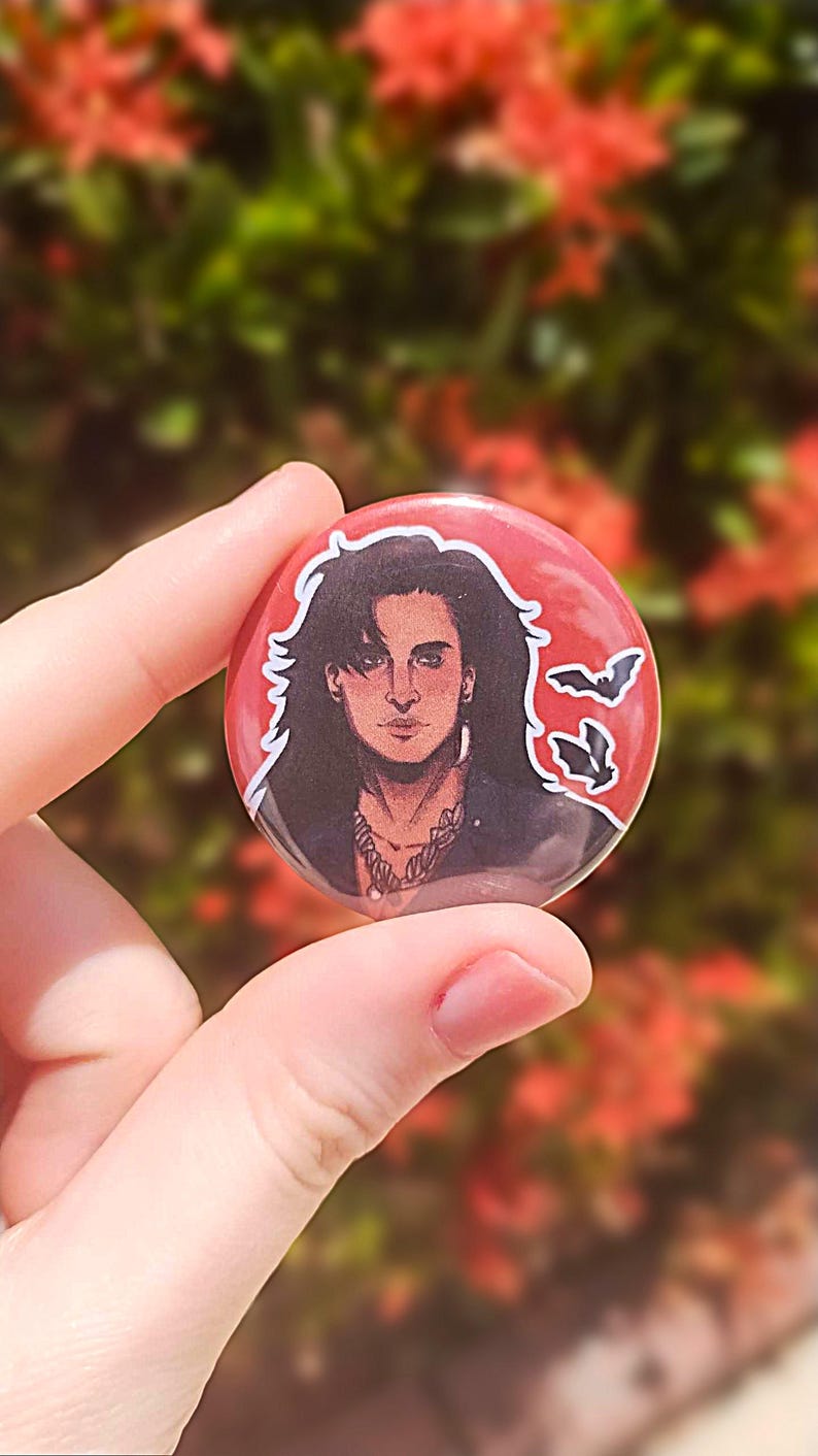 May include: A round button with a red background features a black and white illustration of a man with long black hair. He is wearing a black leather jacket and a silver necklace. Two black bats are flying in the upper right corner of the button.