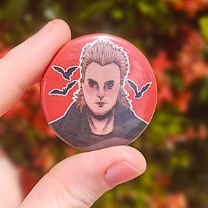 May include: A red button with a black and white illustration of a man with spiky blond hair. The man is wearing a black jacket and a black shirt. There are three black bats flying in the background.