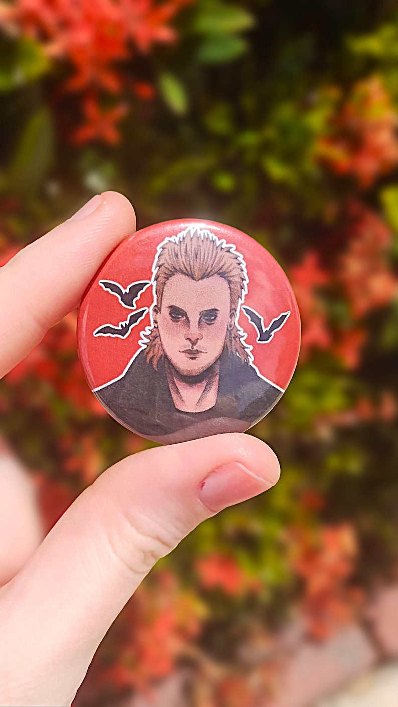 May include: A red button with a black and white illustration of a man with spiky blond hair. The man is wearing a black jacket and a black shirt. There are three black bats flying in the background.