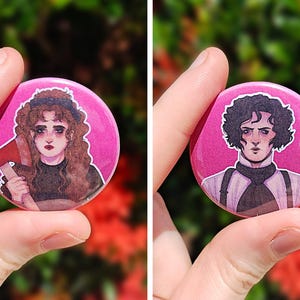 May include: Two round, pink buttons featuring illustrations of characters. One button depicts a woman with an axe, the other a man with dark hair. Both buttons have a white border and are held up by a hand.