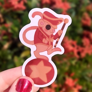 May include: A red and white sticker of a cartoon mouse wearing a red and gold uniform and a hat, balancing on a red ball with a yellow star. The mouse is holding a red and white striped stick.
