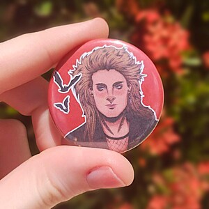 May include: A red button with a black and white illustration of a person with long hair and a dark jacket. The person has a serious expression and is surrounded by bats.