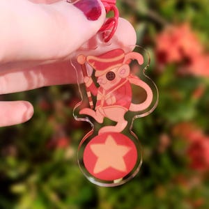 May include: A red and white acrylic keychain featuring a cartoon mouse wearing a red and white uniform and a hat, standing on a red and white star. The mouse is holding a red and white baton.