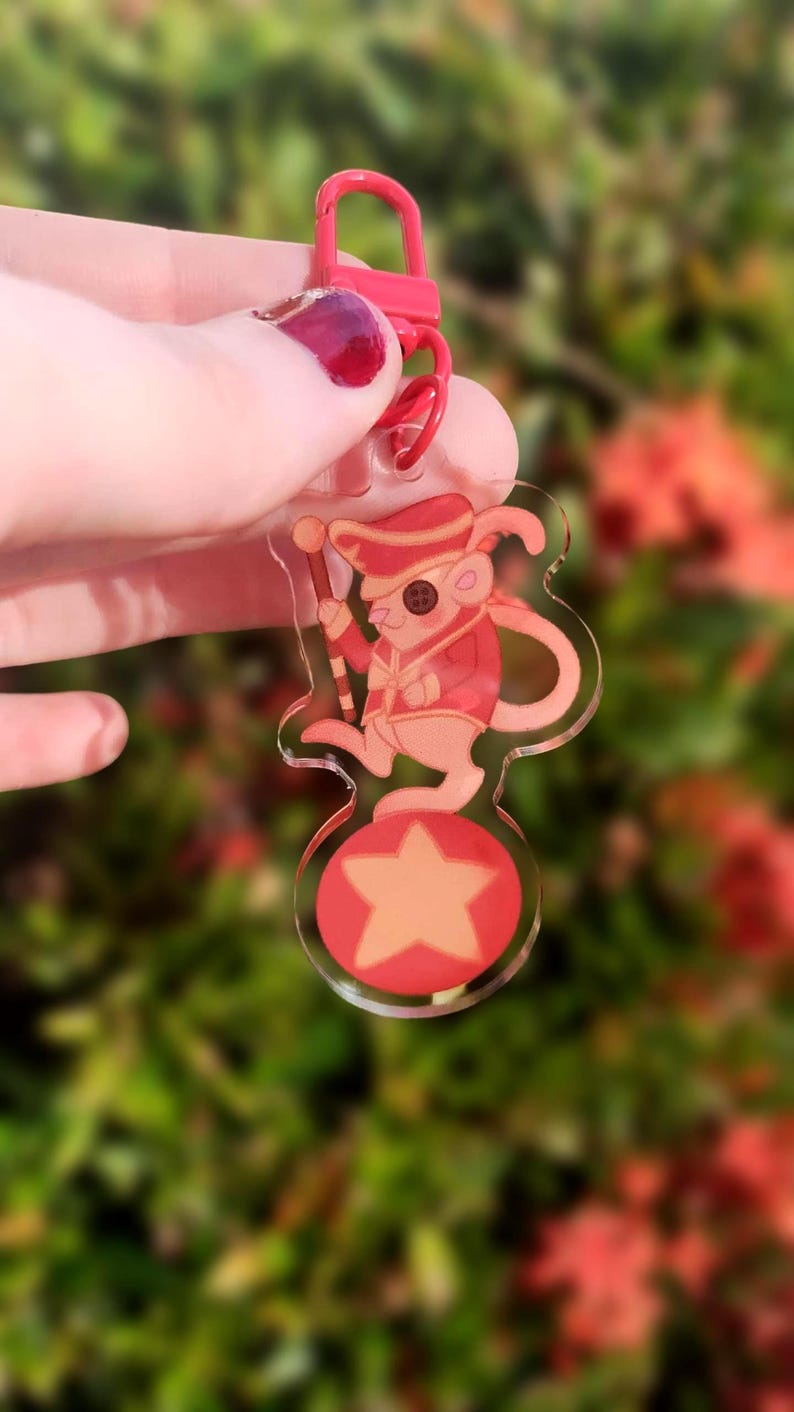 May include: A red and white acrylic keychain featuring a cartoon mouse wearing a red and white uniform and a hat, standing on a red and white star. The mouse is holding a red and white baton.