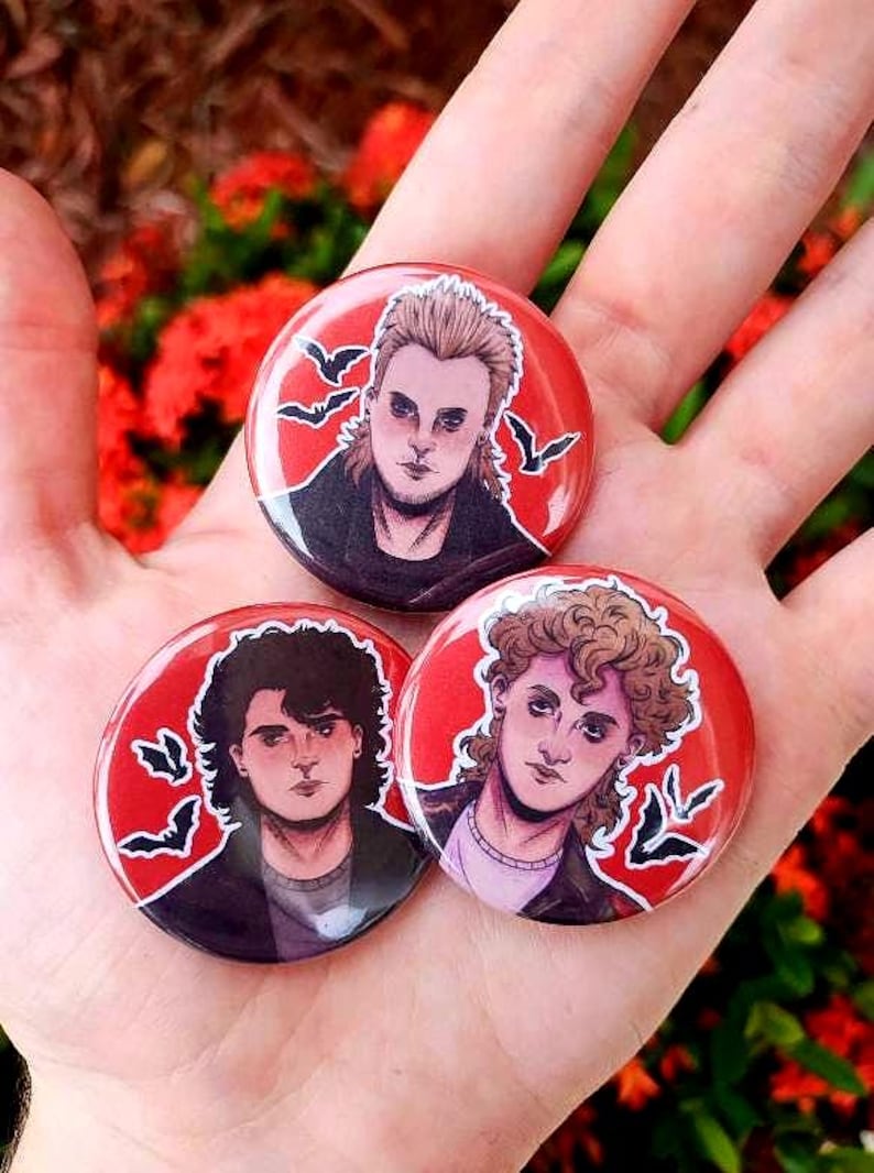 May include: Three round button pins with red backgrounds and black bats. Each pin features a different character from the 1980s movie The Lost Boys. The characters are illustrated in a cartoon style.