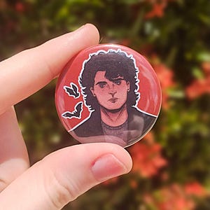 May include: A red button with a black and white illustration of a person with dark hair and a serious expression. The person is wearing a black jacket and there are three white bats in the background.