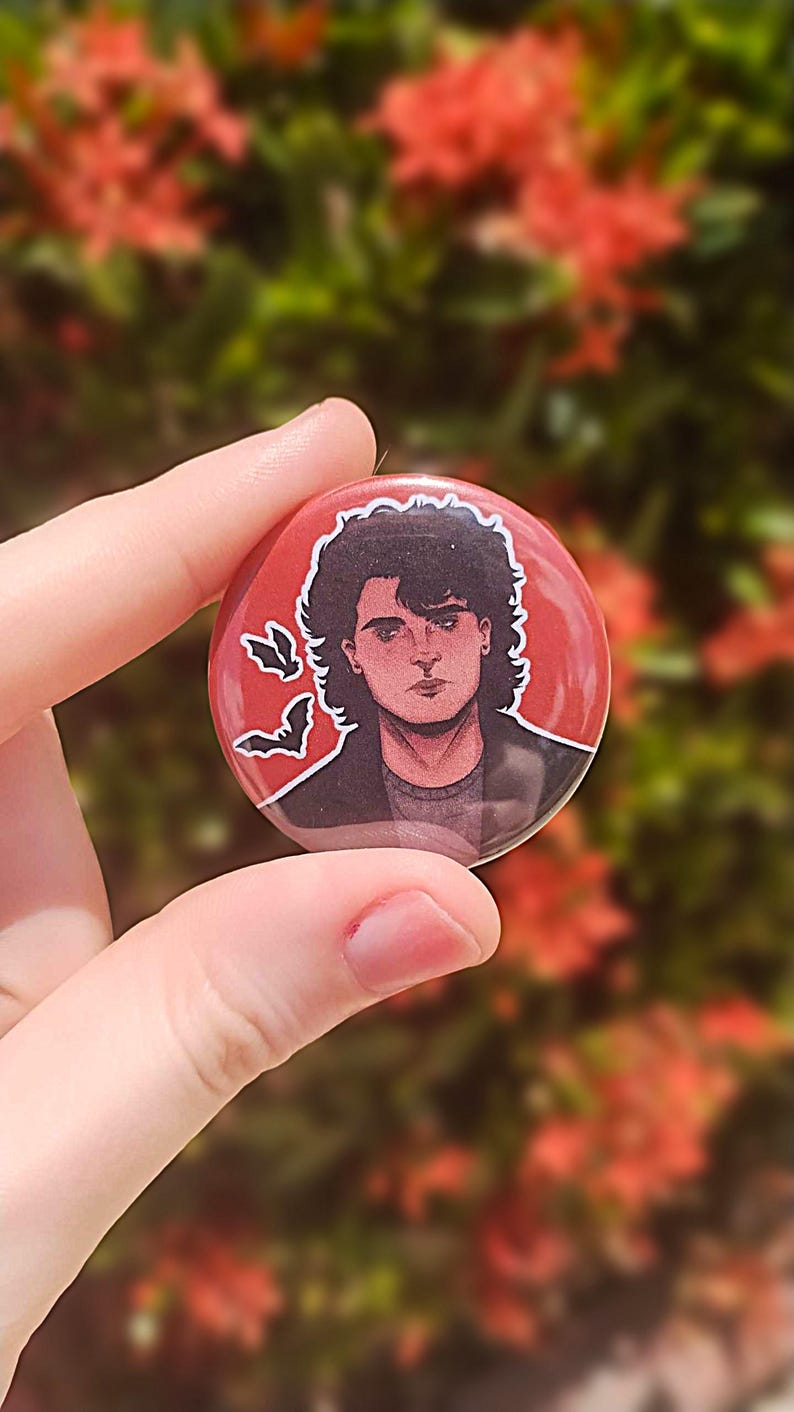 May include: A red button with a black and white illustration of a person with dark hair and a serious expression. The person is wearing a black jacket and there are three white bats in the background.