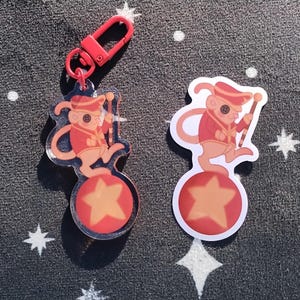 May include: A red and white cartoon mouse wearing a red and gold uniform and a red hat, balancing on a red and yellow ball with a yellow star. The mouse is holding a red and white baton. The image is a keychain charm.