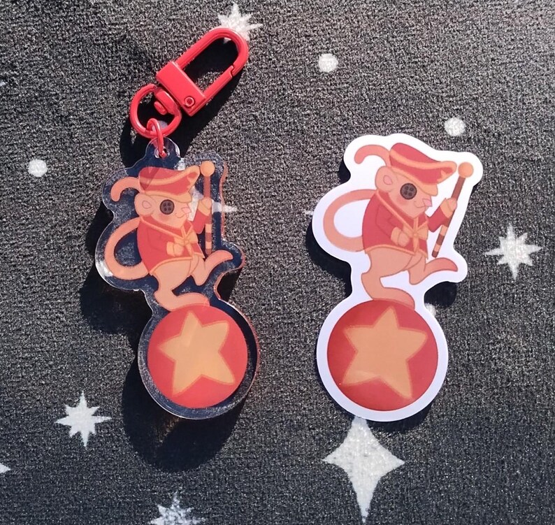 May include: A red and white cartoon mouse wearing a red and gold uniform and a red hat, balancing on a red and yellow ball with a yellow star. The mouse is holding a red and white baton. The image is a keychain charm.