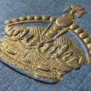 Puede incluir: Primer plano de un emblema dorado y texturizado sobre tela azul. El emblema presenta un diseño detallado con forma de corona y una serie de pequeños acentos redondos. La tela tiene una textura sutil y tejida.