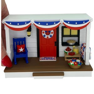May include: A miniature patriotic porch scene with a red door, blue rocking chair, and a wreath. The porch is decorated with red, white, and blue bunting and a welcome mat that says "Happy 4th of July!"