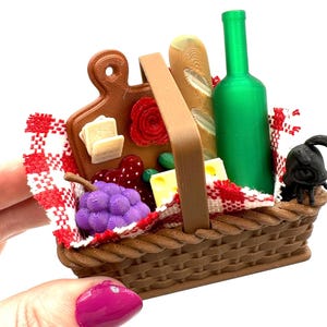 May include: A miniature picnic basket filled with food items. The basket contains a cutting board with cheese and a rose, grapes, a baguette, a green bottle, and a small black figure. A red and white checkered cloth lines the basket.