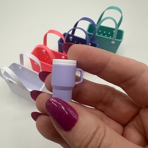 May include: A miniature lavender tumbler with a white lid and handle is held in front of a row of colourful miniature tote bags. The bags are in shades of white, pink, purple, and teal. The tumbler is approximately 2.5 cm tall.