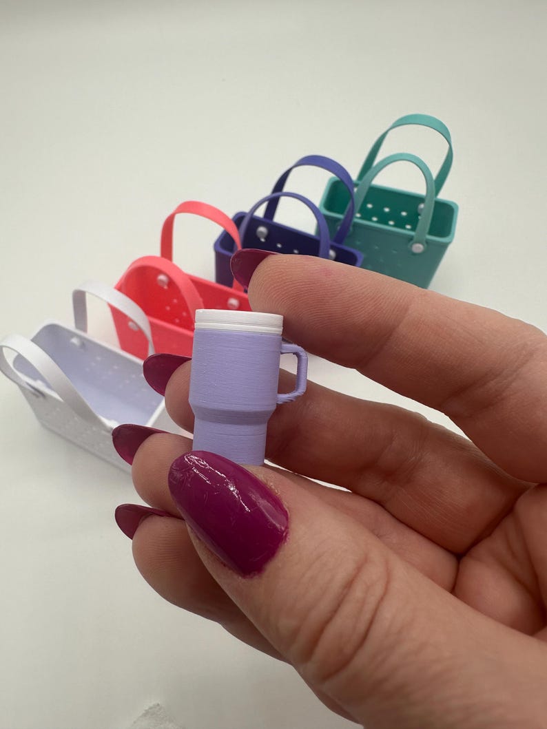 May include: A miniature lavender tumbler with a white lid and handle is held in front of a row of colourful miniature tote bags. The bags are in shades of white, pink, purple, and teal. The tumbler is approximately 2.5 cm tall.