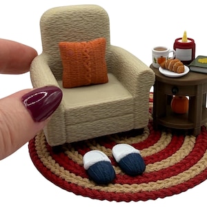 May include: A miniature beige armchair with an orange textured pillow, placed on a round red and beige braided rug. Beside the chair is a small brown side table holding a mug, a croissant, a candle, and a book, with blue slippers nearby.