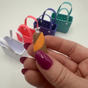 May include: A miniature brown and orange bottle is held in front of a row of colourful miniature baskets. The baskets are in shades of white, pink, purple, and teal. The bottle has a white top.