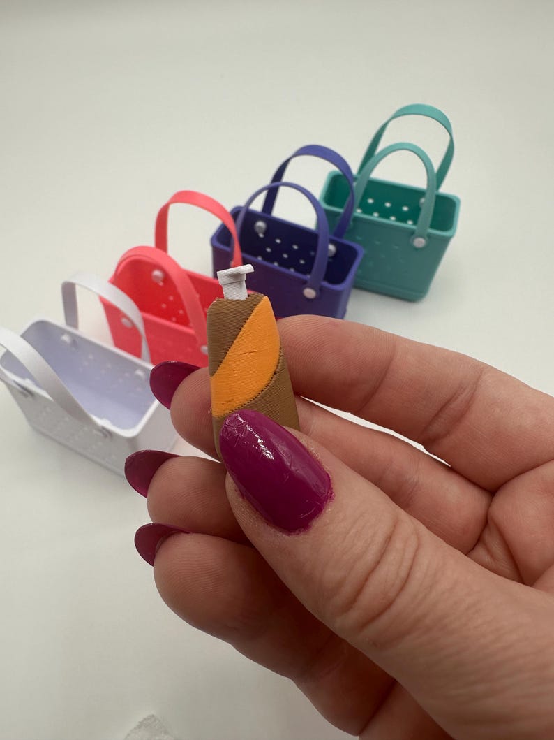 May include: A miniature brown and orange bottle is held in front of a row of colourful miniature baskets. The baskets are in shades of white, pink, purple, and teal. The bottle has a white top.