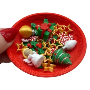May include: A red bowl filled with miniature Christmas ornaments. The ornaments include gold stars, green gift boxes with red bows, white and red peppermint candies, a gold angel, and a green Christmas tree.