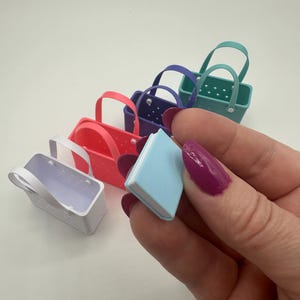 May include: A selection of small, brightly coloured tote bags in red, purple, teal, and white, alongside a tiny blue book. The bags feature handles and a rectangular design. The book is held by a person with purple nail polish.