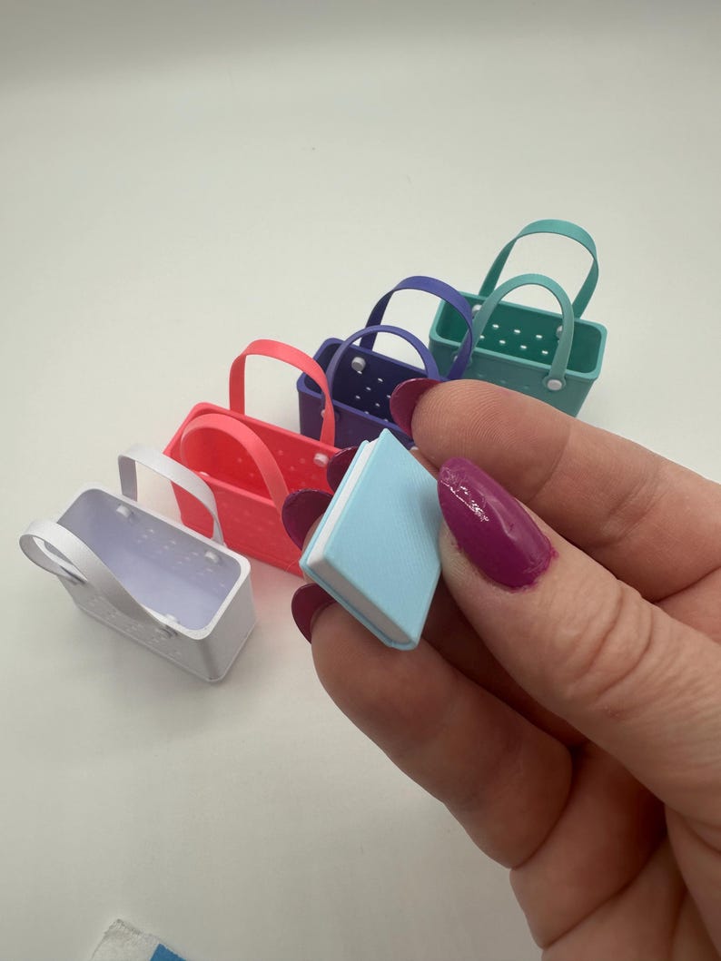 May include: A selection of small, brightly coloured tote bags in red, purple, teal, and white, alongside a tiny blue book. The bags feature handles and a rectangular design. The book is held by a person with purple nail polish.
