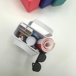 May include: A white tote bag containing a rolled red and white towel, sunglasses, and other small items. Several colourful containers with rolled towels are in the background. The tote bag has a white handle.