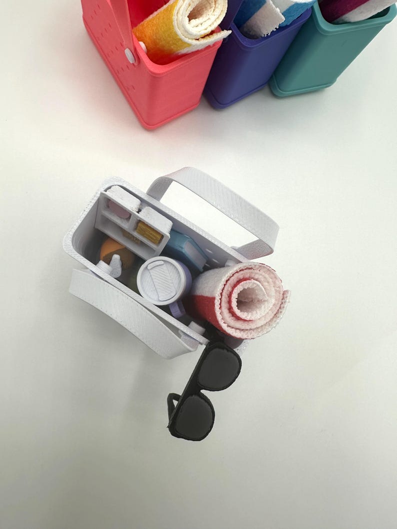 May include: A white tote bag containing a rolled red and white towel, sunglasses, and other small items. Several colourful containers with rolled towels are in the background. The tote bag has a white handle.