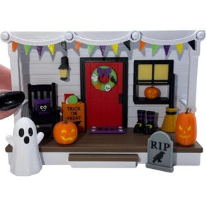 May include: A miniature Halloween porch scene featuring a white house facade with a red door, a green wreath, a black rocking chair, a trick or treat bag, pumpkins, a ghost figurine, a tombstone with RIP text, and colorful bunting.
