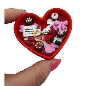 May include: A red heart-shaped container filled with miniature Valentine's Day themed decorations. The decorations include chocolates, bows, hearts, a letter, and an arrow. The container is held in a person's hand.