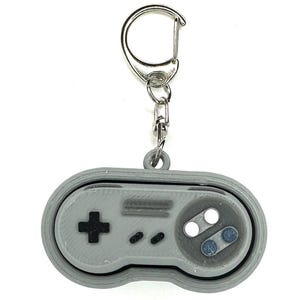May include: A gray Super Nintendo controller keychain with a silver key ring. The controller has a black plus button, two black buttons, and two gray buttons.