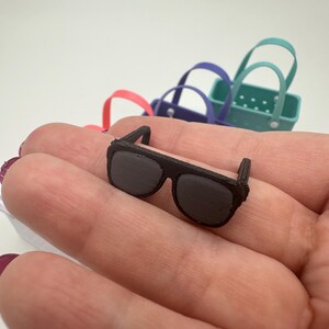 May include: A miniature pair of dark brown sunglasses with a rectangular frame and dark lenses. The sunglasses are resting on a person's palm, with several small, colourful tote bags in the background.