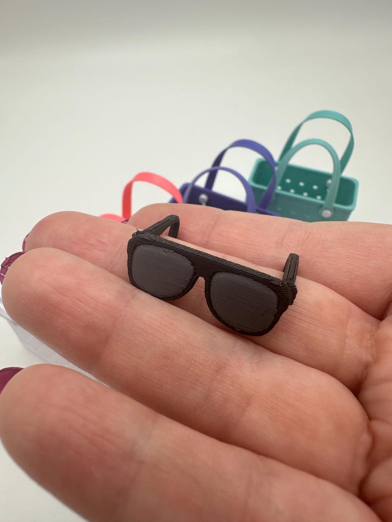 May include: A miniature pair of dark brown sunglasses with a rectangular frame and dark lenses. The sunglasses are resting on a person's palm, with several small, colourful tote bags in the background.