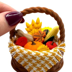 May include: A miniature brown basket with a braided handle containing small plastic autumn-themed items including a pumpkin, corn, leaves, and a gourd, draped with a yellow and white houndstooth patterned fabric cloth with fringed edges.