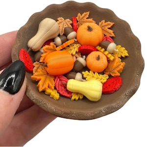 May include: A brown bowl filled with miniature autumn-themed decorations. The bowl contains orange pumpkins, yellow and red leaves, acorns, and gourds. The decorations are in various shapes and sizes, creating a festive fall display.