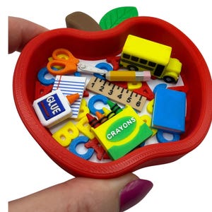 May include: A red apple-shaped container filled with miniature school supply erasers including a yellow school bus, a ruler, a glue bottle, a blue book, scissors, a box labeled CRAYONS, and various colorful alphabet letters scattered inside.
