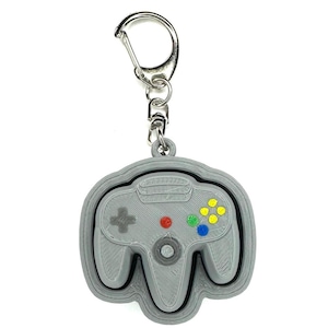 May include: A gray keychain featuring a 3D-printed design of a classic video game controller. The controller has colorful buttons and a silver metal clasp for attaching to keys or a bag.