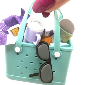 May include: A miniature turquoise tote bag filled with picnic items, including a purple towel, a mug, a cupcake, and a bottle. Black sunglasses hang from the side of the bag. The bag has a handle and white accents.