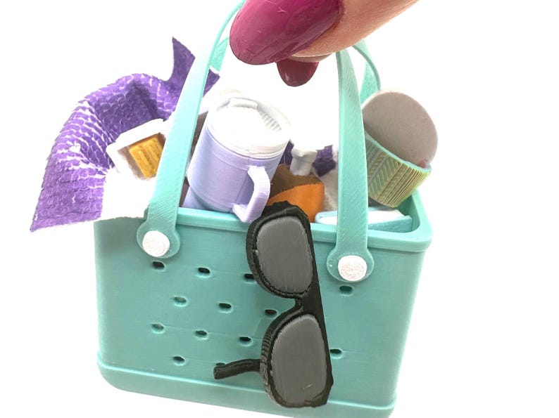 May include: A miniature turquoise tote bag filled with picnic items, including a purple towel, a mug, a cupcake, and a bottle. Black sunglasses hang from the side of the bag. The bag has a handle and white accents.