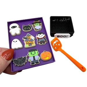 May include: A purple rectangular tray holding eight small Halloween-themed figures including a mummy, black cat, witch, ghost, haunted house, vampire, bat, Frankenstein's monster, and pumpkin. Beside it is a black basket labeled BOO! and an orange spatula.