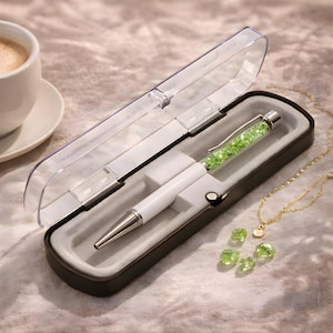 May include: A white pen with a silver tip and a green crystal-filled barrel, presented in a black case with a clear lid. A gold necklace with green gemstones and a small pendant is also visible.